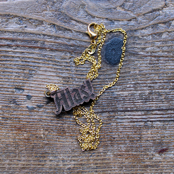 Walnut wood necklace in the shape of text on a gold-coloured metal chain, sitting on a wooden plank.