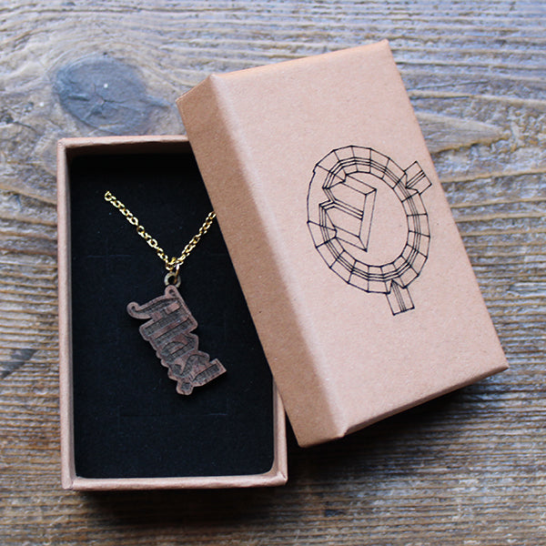 Walnut wood necklace in the shape of text on a gold-coloured metal chain, sitting in a kraft paper gift box