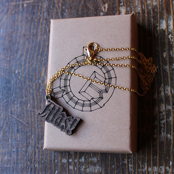 Walnut wood necklace in the shape of text on a gold-coloured metal chain, sitting on a kraft paper gift box