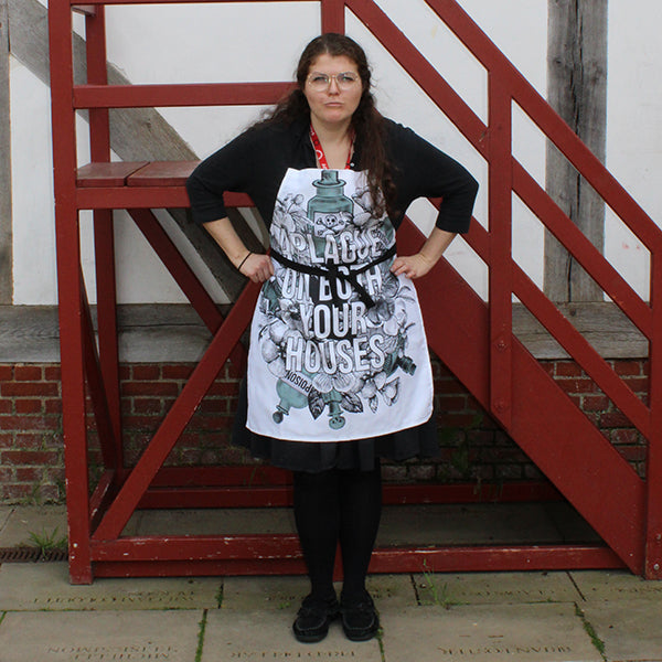 White cotton apron with black neck loop and ties, printed with an image celebrating Shakespeare play, Romeo & Juliet. A background of black and white flowers and green poison bottles with a quote fro the play, "a plague on both you house" printed in white capital letters.