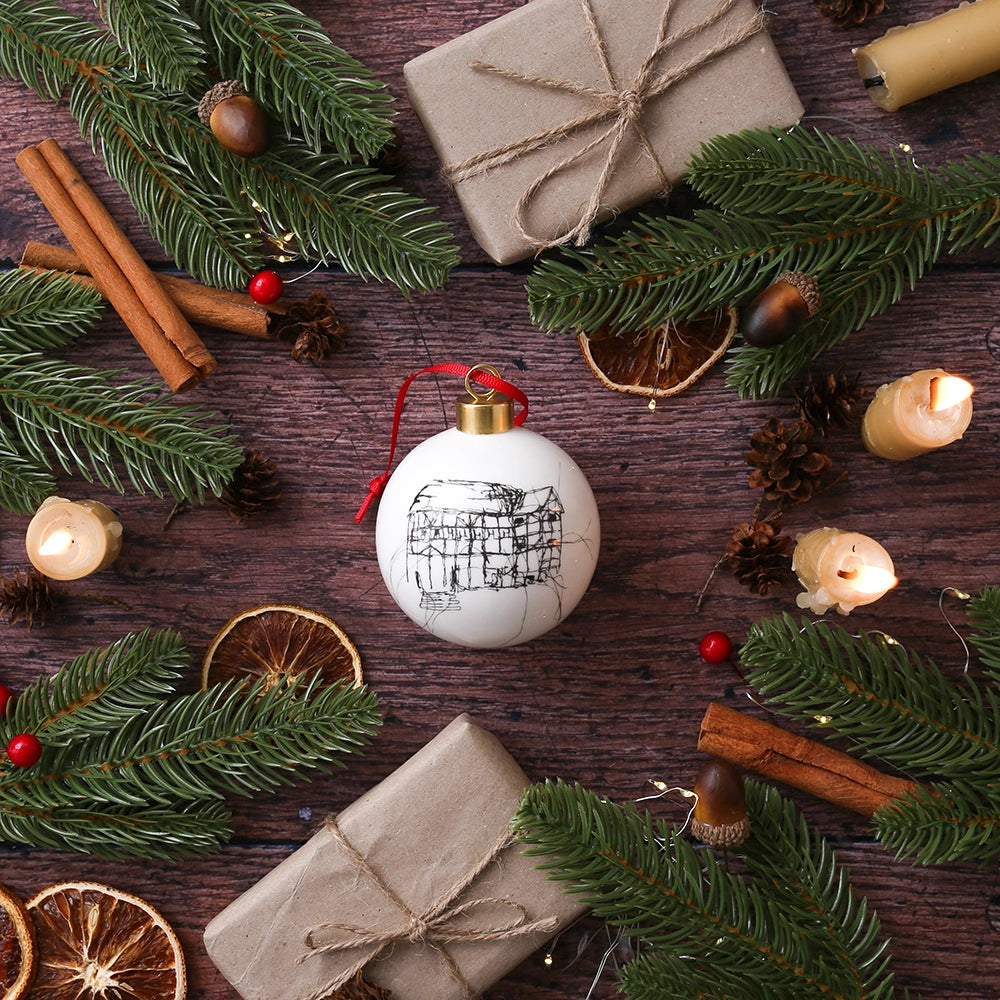 White glass bauble with black etched drawing of Shakespeare's Globe and gold top, with red tied ribbon, on wooden background with Christmas greenery, candles, orange slices and wrapped brown paper packages