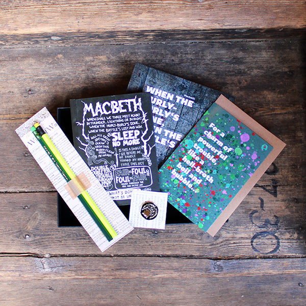 Black box containing pencils on a backing card, a journal with white print, a green greetings card, a grey print and a black and gold pin badge.