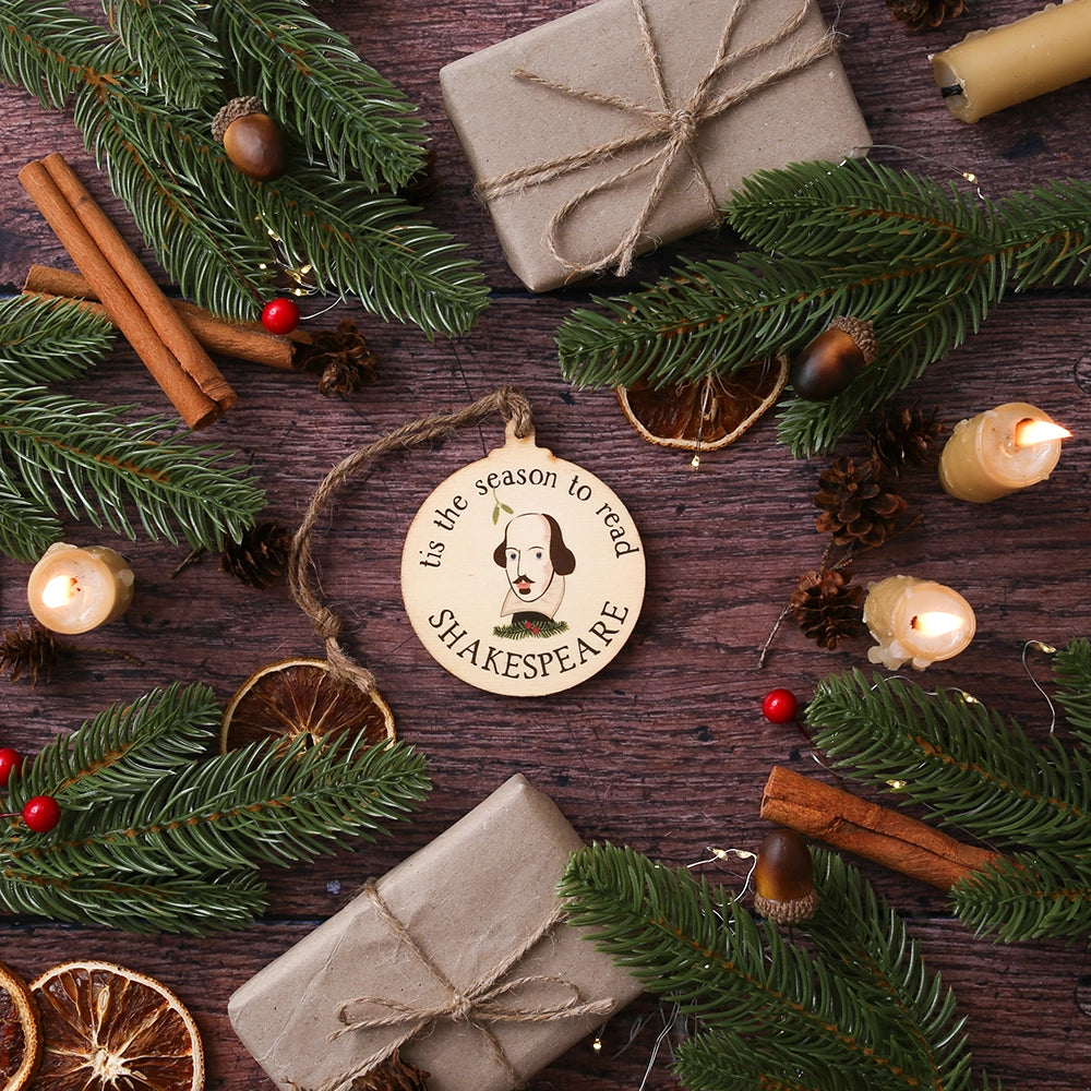Round, flat, wooden Christmas decoration with Shakespeare's face in centre surrounded by 'tis the season to read' text., on wooden background with Christmas greenery