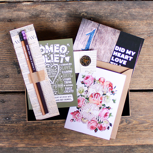 A kraft box containing pencils on a backing card, a green journal with white print, a black and gold pin badge, a purple and brown print and a floral greetings card