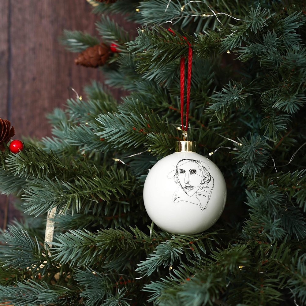 White fine china hanging bauble with a gold ferule and red ribbon. On the bauble is a portrait of William Shakespeare based on an embroidery. The portrait is made up of black lines and has a scribbled look, on a Christmas tree