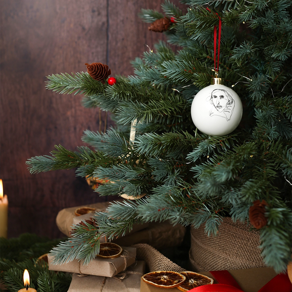 White fine china hanging bauble with a gold ferule and red ribbon. On the bauble is a portrait of William Shakespeare based on an embroidery. The portrait is made up of black lines and has a scribbled look, on a Christmas tree