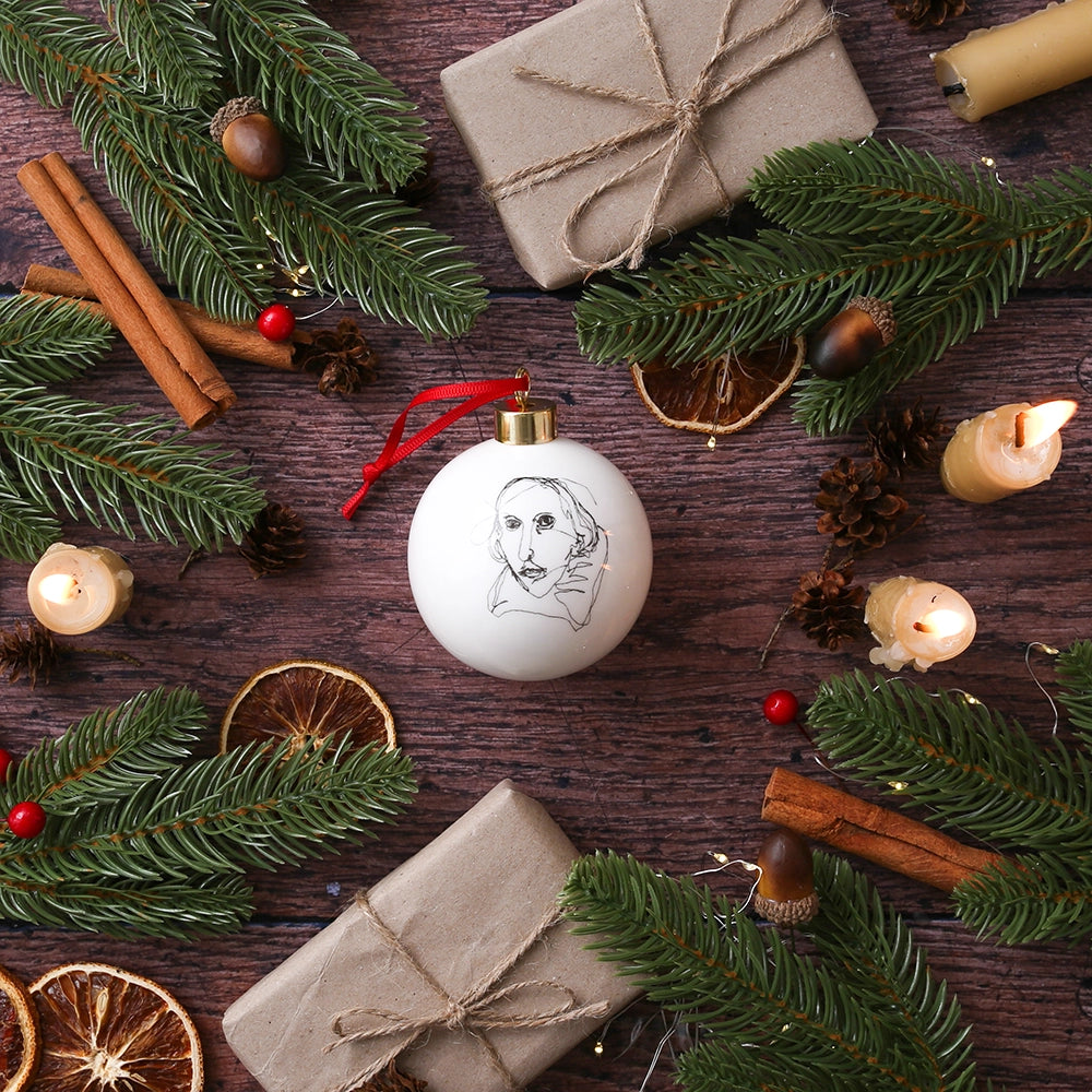 White fine china hanging bauble with a gold ferule and red ribbon. On the bauble is a portrait of William Shakespeare based on an embroidery. The portrait is made up of black lines and has a scribbled look.