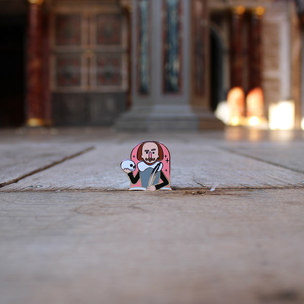 Cartoon metal pin badge of Shakespeare's torso with one hand holding a quill and the other a skull