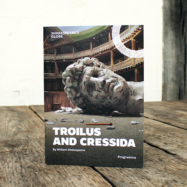 Theatre programme for 'Troilus and Cressida' by William Shakespeare on a wooden surface with a background of Shakespeare's Globe theatre.