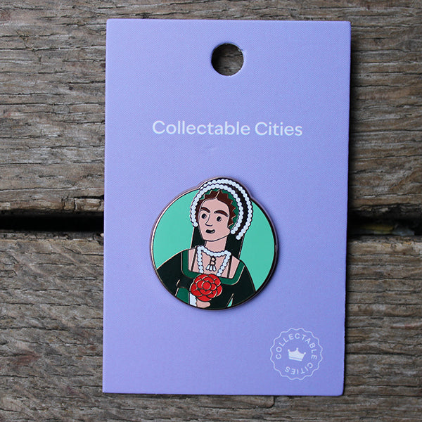 Teal coloured circular pinbadge, with cartoon graphic of Anne Boleyn in the centre, on purple card backing, sitting on wooden stage