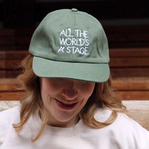 Khaki green six-panel baseball cap with a pre-curved peak. Embroidered on the front of the cap is a quote from Shakespeare play, As You Like It (all the world's a stage) in white hand-drawn capital letters.