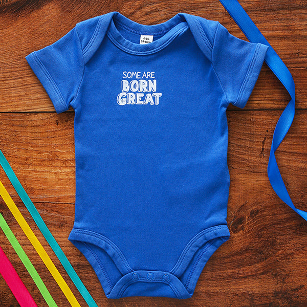 Royal blue cotton baby grow with short sleeve and white graphic text on centre front, on wooden background with coloured ribbons