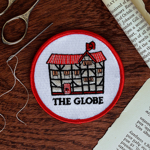 Embroidered patch of 'The Globe' theater on a wooden surface with sewing tools.