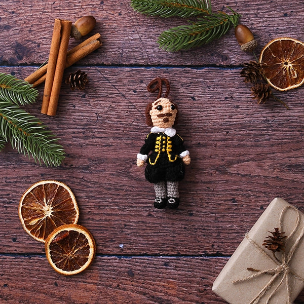 Crocheted Shakespeare decoration wearing a black doublet and hose with grey stockings on wooden plank background with cinnamon stick, dried orange slices, wrapped brown package and Christmas greenery