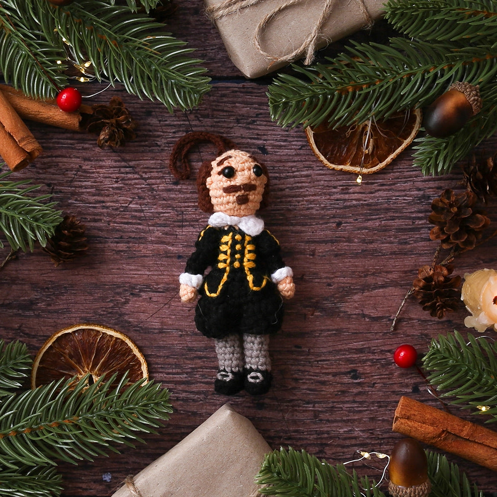 Crocheted Shakespeare decoration wearing a black doublet and hose with grey stockings on wooden plank background with cinnamon stick, dried orange slices, wrapped brown package and Christmas greenery