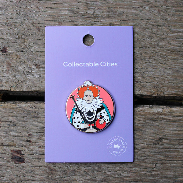 Circular pink pinbadge with graphic of cartoon Elizabeth I, sitting on purple card backing, lying on wooden stage