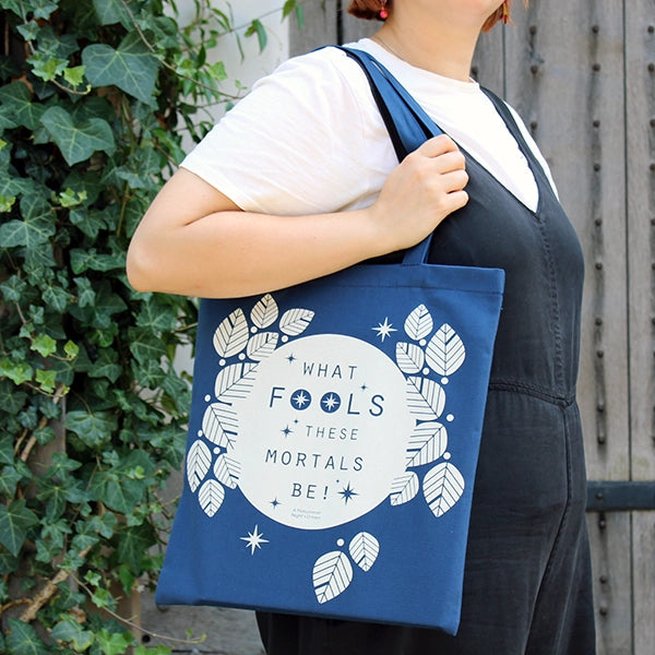 A  cotton book bag for Shakespeare's Globe. The bag is midnight blue with mid-length matching handles. Printed on the bag in white is a design consisting of a white circle (to represent the full moon) surrounded by stylised white leaves and stars. Across the face of the moon is a quote from Shakespeare play, A Midsummer Night's Dream (What fools these mortals be!) in midnight blue. The lettering is slim capitals.