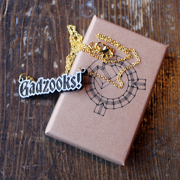 Walnut wood necklace in the shape of text on a gold-coloured metal chain, sitting on a kraft paper gift box