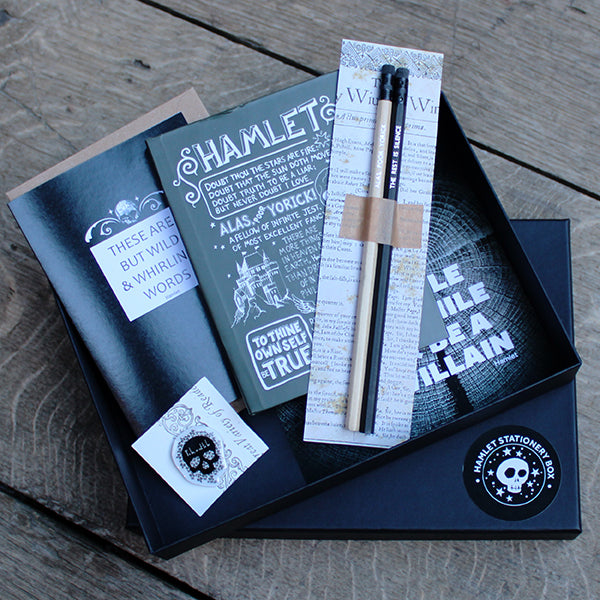 A black gift box containing pencils on a backing card, a grey journal with white text, a black and white print, a black greetings card and a black and white pin badge