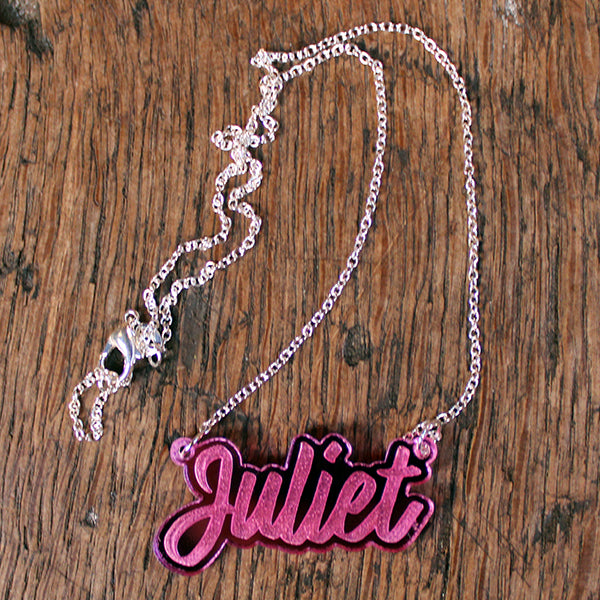 Pink perspex necklace in script text with the word 'Juliet' spelled out on silver chain, lying on wooden background