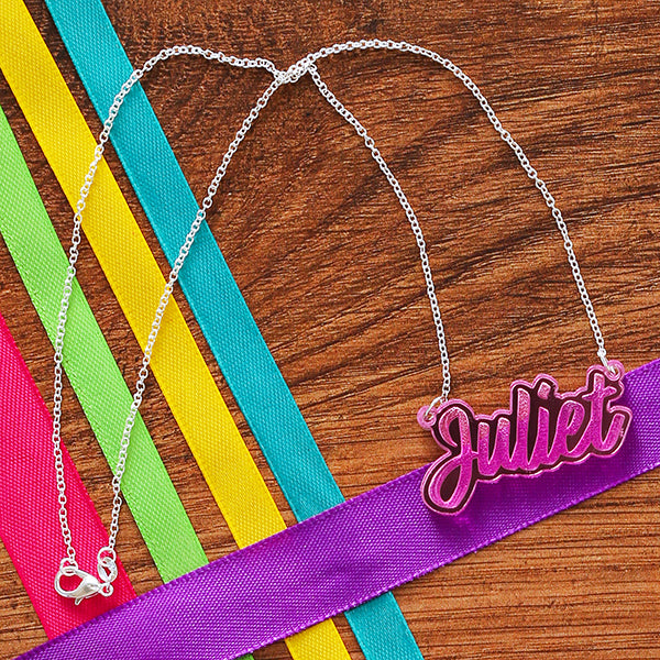 Pink perspex necklace in script text with the word 'Juliet' spelled out on silver chain, lying on wooden background with multi-coloured ribbons