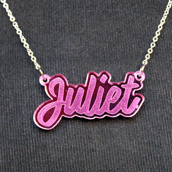 Pink perspex necklace in script text with the word 'Juliet' spelled out on silver chain, lying on black fabric background