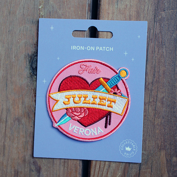 Pink circular sew on patch, with tattoo style graphics, red heart and dagger, on purple backing card, sitting on wooden surface