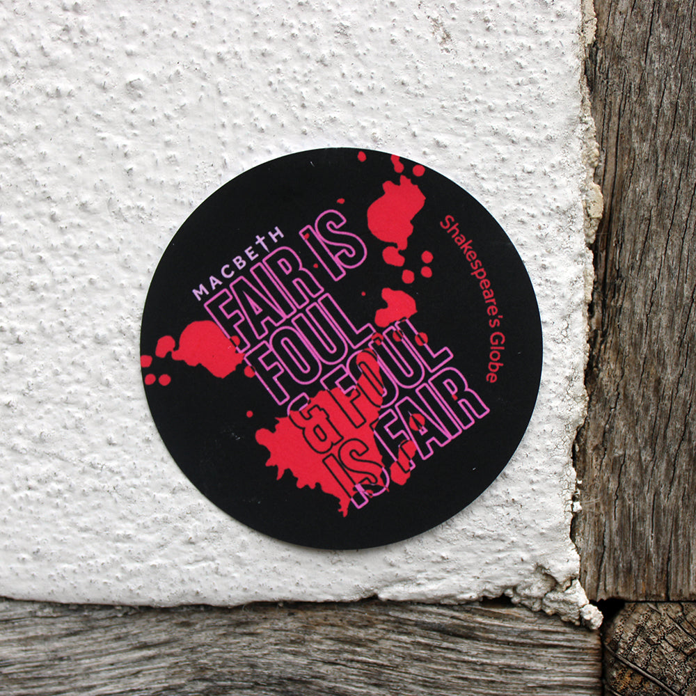 Black circular sticker with pink outlined graphic text across it, covered in bold red blood splatter design, stuck on a white textured wall