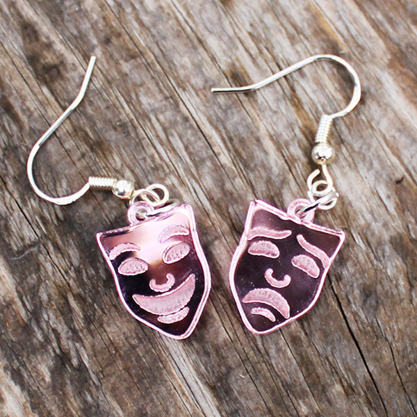 Pair of rose gold coloured mask-shaped earrings on a wooden surface
