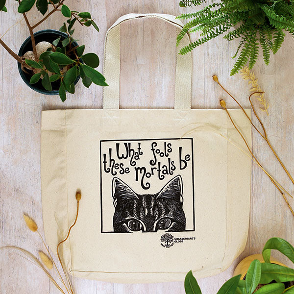 Beige tote bag with black cat illustration and text on a wooden surface with plants.