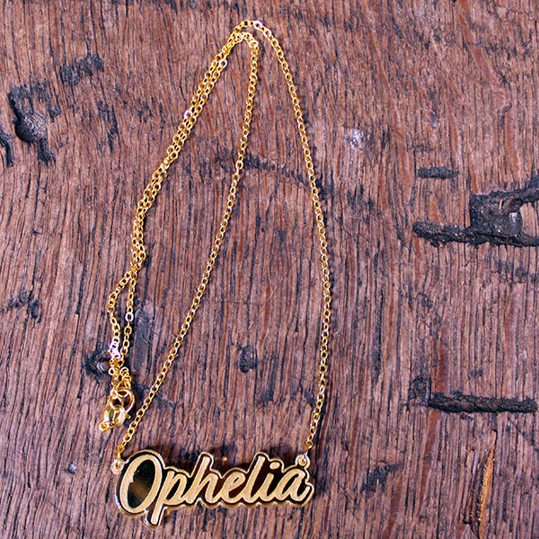 Gold perspex necklace in script text with the word 'Ophelia' spelled out on gold chain, lying on wooden background