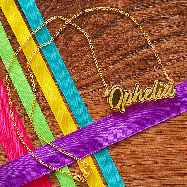 Gold perspex necklace in script text with the word 'Ophelia' spelled out on gold chain, lying on wooden background with coloured ribbons