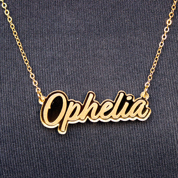 Gold perspex necklace in script text with the word 'Ophelia' spelled out on gold chain, lying on black fabric background