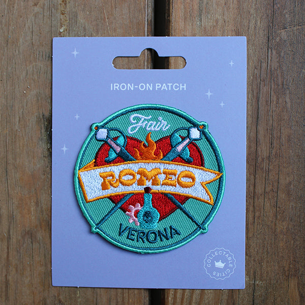 Teal coloured circular patch with red heart and daggers, on purple card backing, sitting on wood surface