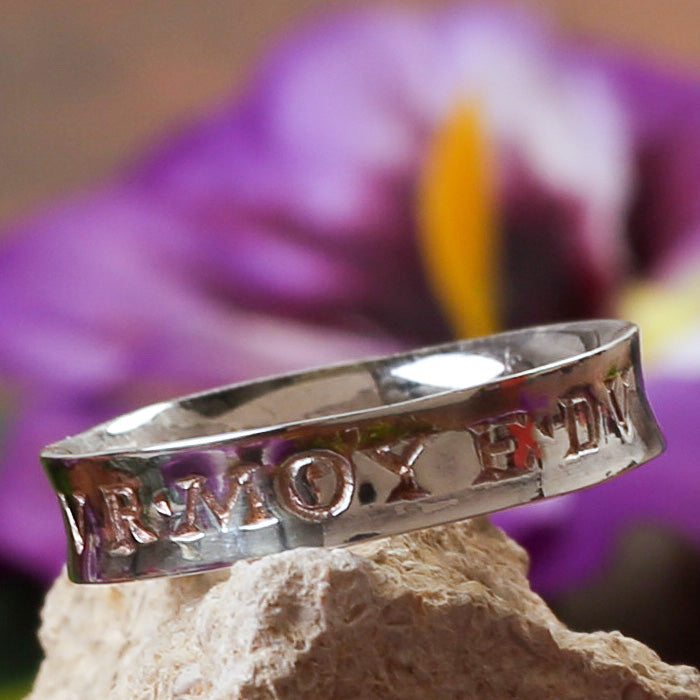 A silver ring with an engraved message and decorative elements, displayed on a rock with a flower in the background.