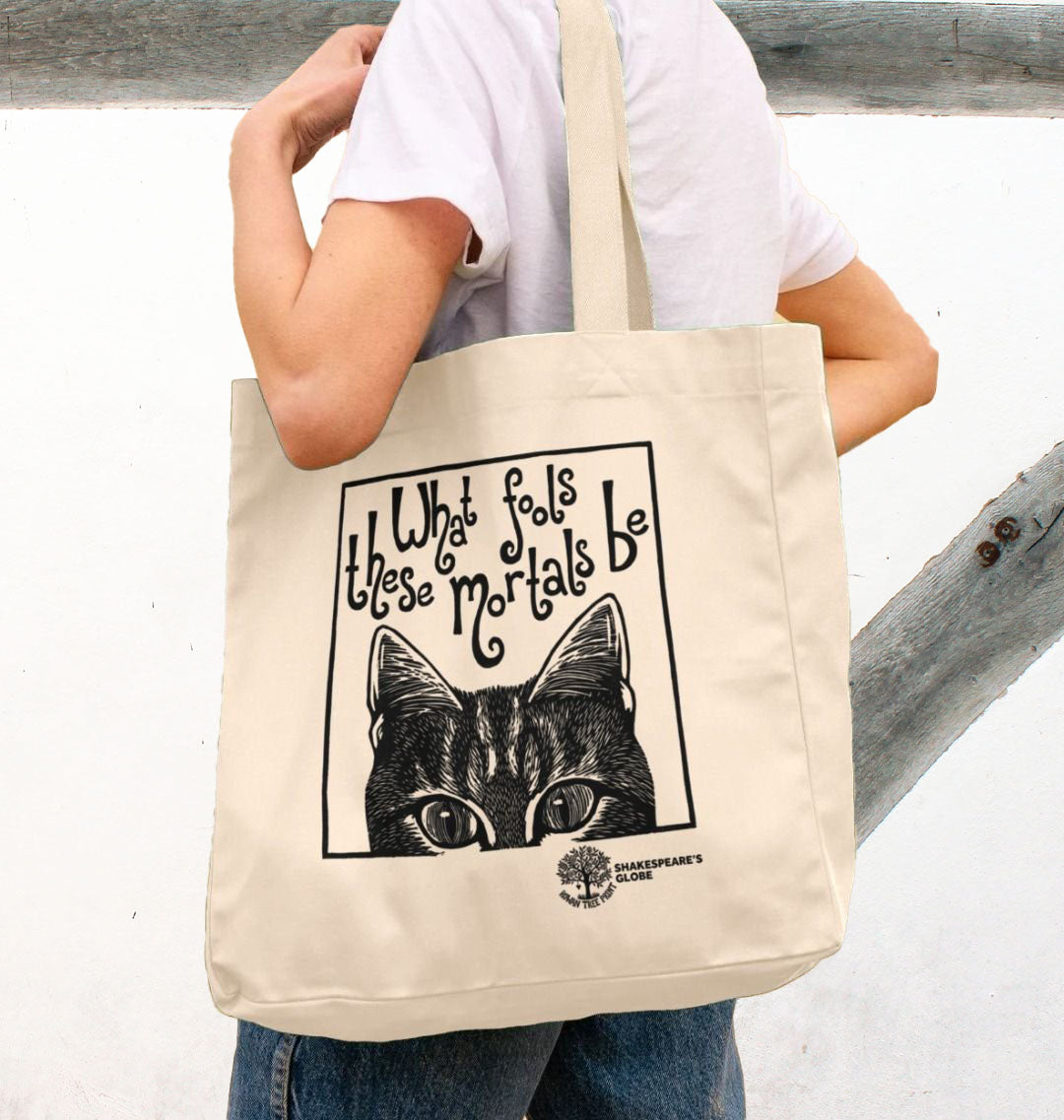 Person holding a tote bag with a cat illustration and text, standing against a limewashed wall.
