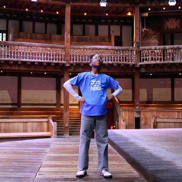 Young girl with glasses standing with hands on hips on Globe stage with grey jeans and royal blue t-shirt with white text