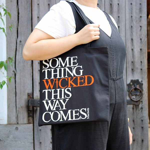 Black cotton bag with mid-length black handles printed with a quote from Shakespeare play, Macbeth (Something wicked this way comes). The lettering is bold serifed capital letters which are slightly distressed printed in white, except for 'WICKED) which is printed in orange. The 'I' of wicked is replaced by a dagger.