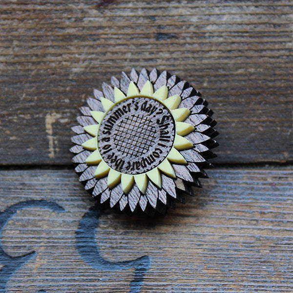walnut wood and cream acrylic brooch in the shape of a stylised flower with text engraved around the centre