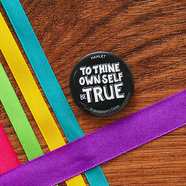 38 mm diametre grey button badge with a quote from Shakespeare play, Hamlet (to thine own self be true) printed in white, hand-drawn, capital letters, on wooden background with coloured ribbons