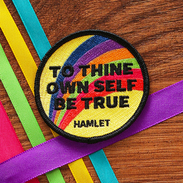 Rainbow yellow patch with black border and bold black text, circular shape, sitting on wood background with coloured ribbons