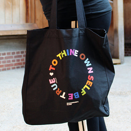 Black cotton tote bag with 2 handles, with circular text graphic showing letters in the colours of the pride flag