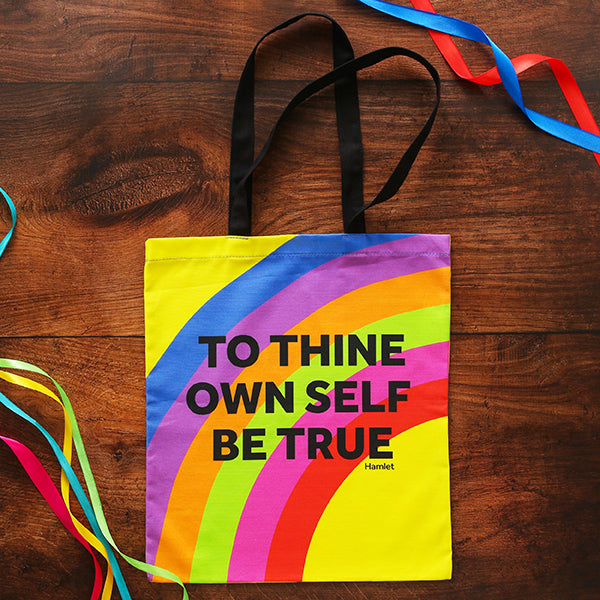 Rainbow coloured tote bag with black handles and text, on wooden background with multi coloured ribbons
