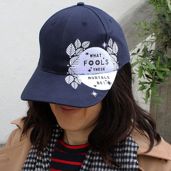 Navy blue six panel baseball cap with an embroidered motif in white across the left side of the cap and peak