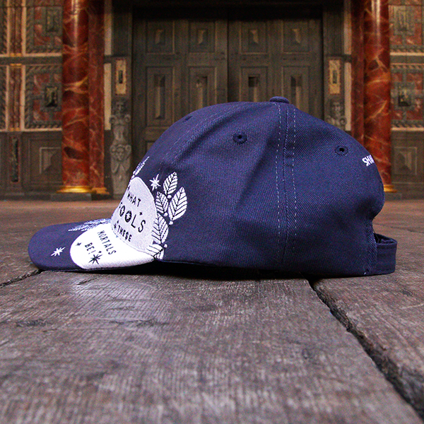 Navy blue six panel baseball cap with an embroidered motif in white across the left side of the cap and peak. The motif consists of the full moon surrounded by stylised leaves and stars. Across the face of the moon is a quote from Shakespeare play, A Midsummer Night's Dream, (what fools these mortals be!) in blue.