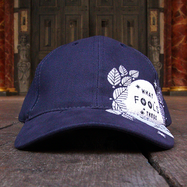Navy blue six panel baseball cap with an embroidered motif in white across the left side of the cap and peak. The motif consists of the full moon surrounded by stylised leaves and stars. Across the face of the moon is a quote from Shakespeare play, A Midsummer Night's Dream, (what fools these mortals be!) in blue.
