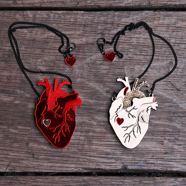 Two Pendants in the shape of an anatomical heart. One pendant is made of birch wood in several layers, giving the appearance of three dimensions. The top layer has channels cut to represent veins and the under layers are textured. A cutout in the shape of a graphic heart sits about halfway down the pendant and is filled in with bright red Persex. The second pendant is made in a similar way using bright red Perspex with a silver Perspex graphic heart cutout. Both pendants are on black chains.