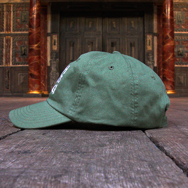 Khaki green six-panel baseball cap with a pre-curved peak. Embroidered on the front of the cap is a quote from Shakespeare play, As You Like It (all the world's a stage) in white hand-drawn capital letters.