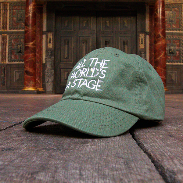 Khaki green six-panel baseball cap with a pre-curved peak. Embroidered on the front of the cap is a quote from Shakespeare play, As You Like It (all the world's a stage) in white hand-drawn capital letters.