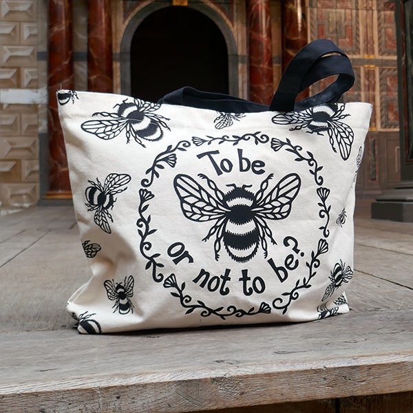 Tote bag made of heavyweight unbleached cotton canvas with mid-length black webbing handles. The bag is printed with a black design adapted from an original print of a bumble bee with a striped body and paneled wings, and a quote from Shakespeare play, Hamlet (To be or not to be?). In a ring around the bee and the quote are flowers and leaves. Outside this main design are a number of smaller bees flying in different directions